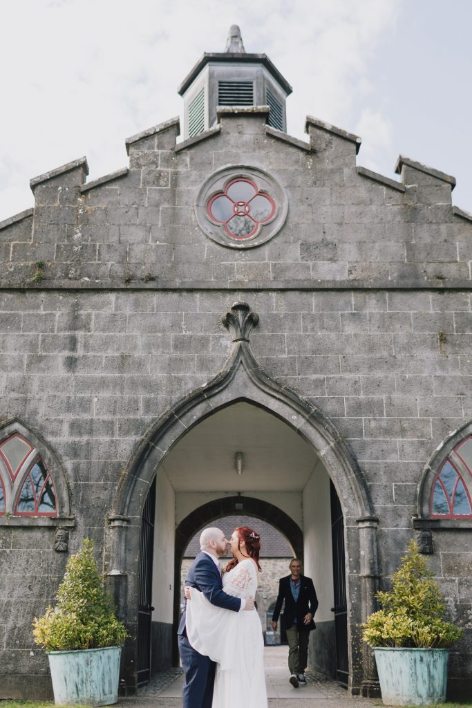 Slane Castle Wedding Couple kissing each other while owner's son Alex Conyngham son of Lord Henry Mountcharles walking past them under the arch